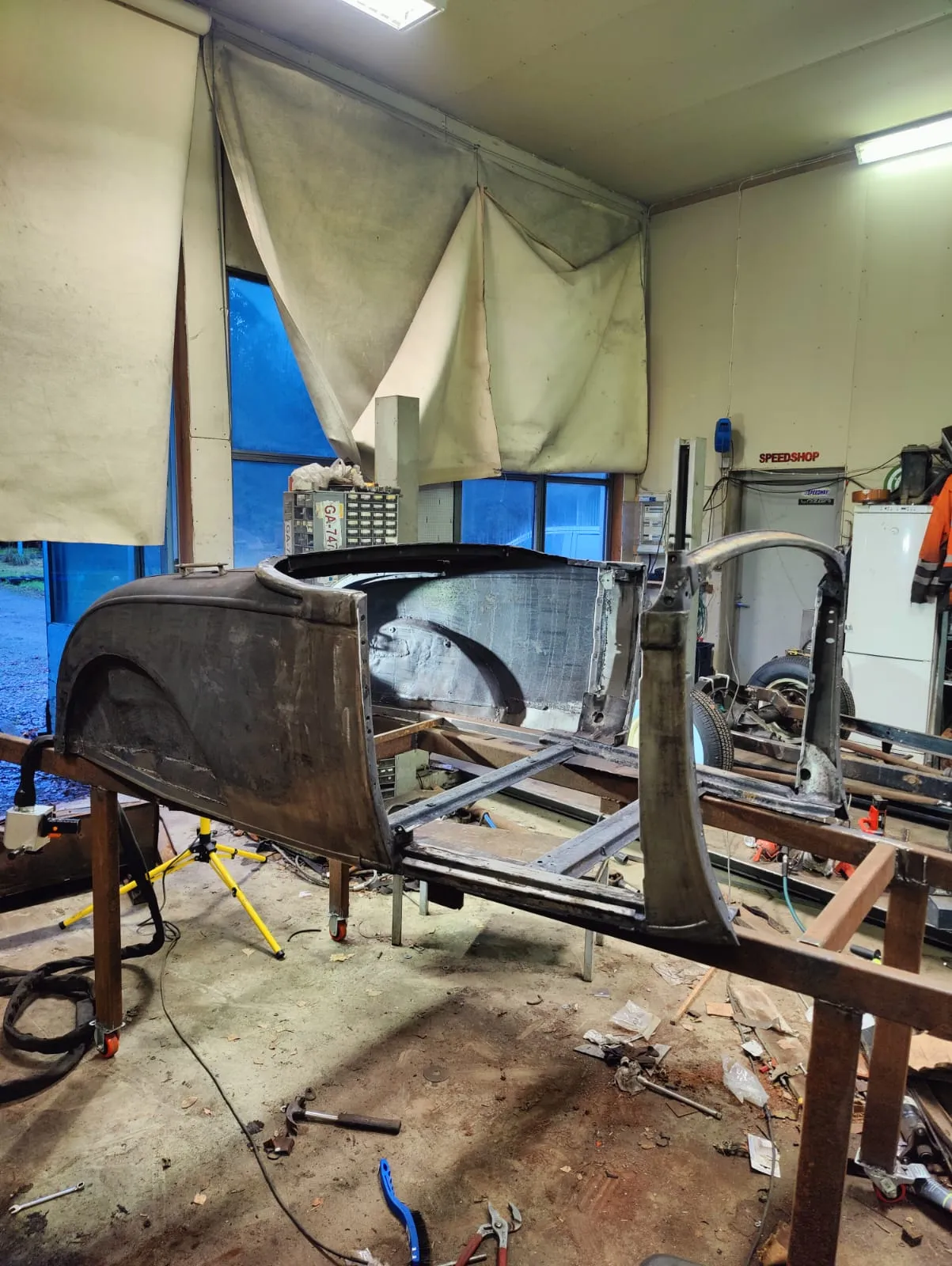 Työpajassa kuvattu vanhan auton korin kunnostusprojekti. Tumma, maalaamaton metallinen auton kori on nostettu ruskealle metallitelineelle. Taustalla on ikkunoita, joista näkyy sininen ulkoilma, sekä valkoisia pressuja ja varastokaappeja. Lattialla on työkaluja, kuten vasara ja pihdit, sekä kaapeleita. Katossa on kirkkaat loisteputkivalot.